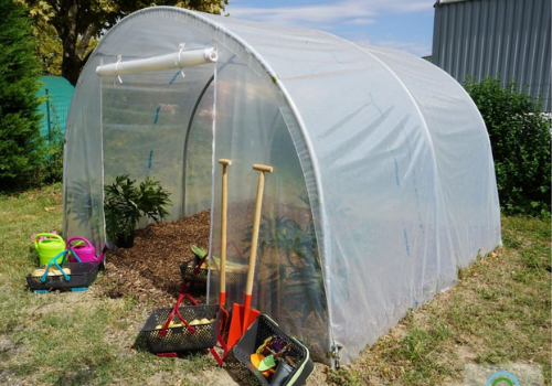 Petite serre tunnel, porte en bâche ouverte avec accessoires de jardinage : panniers, pelles, arrosoirs, ...